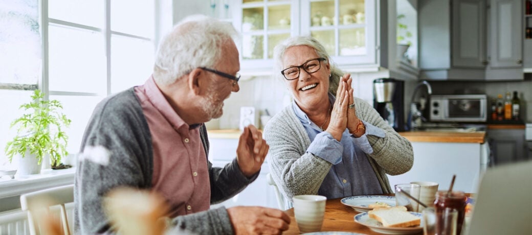 Older man and woman sitting together in the kitchen while having a conversation and laughing.